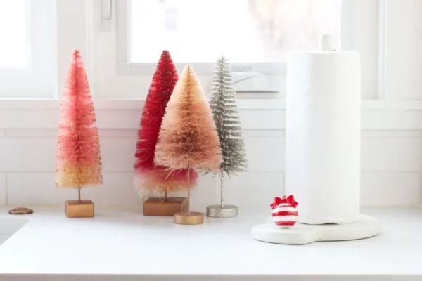 Nora Fleming Mini Deck the Halls Red Christmas ornament attached to a paper towel holder beside decorative trees